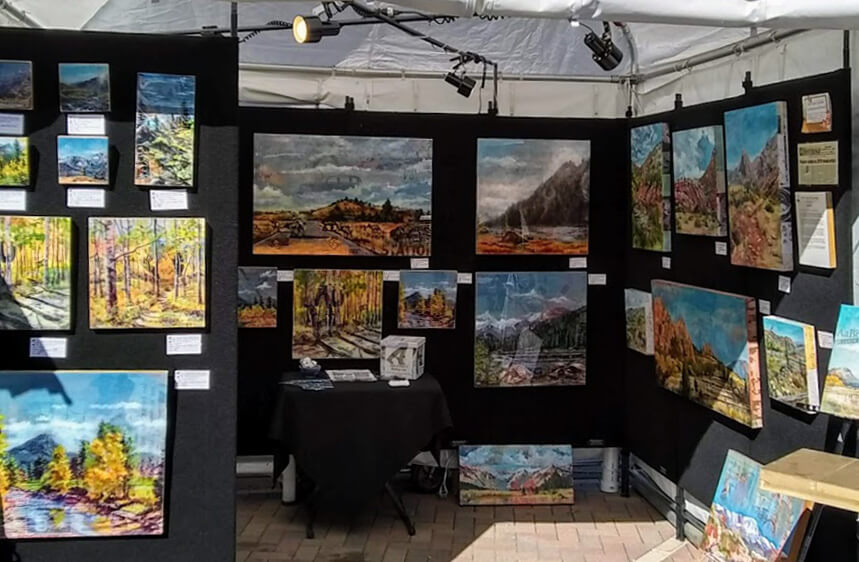 Artist booth in outdoor art show with paintings hung on a black background