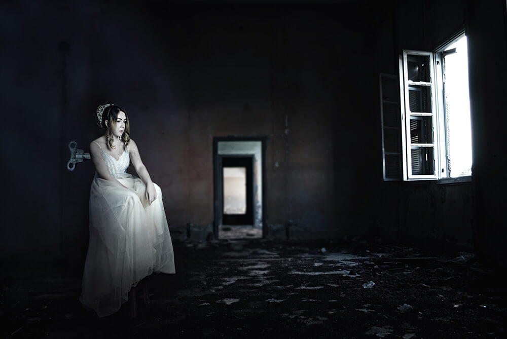 Photo illustration of a woman in a wedding dress and tiara sitting on a stool in a dark room. A wind-up knob is extending from her back. 
